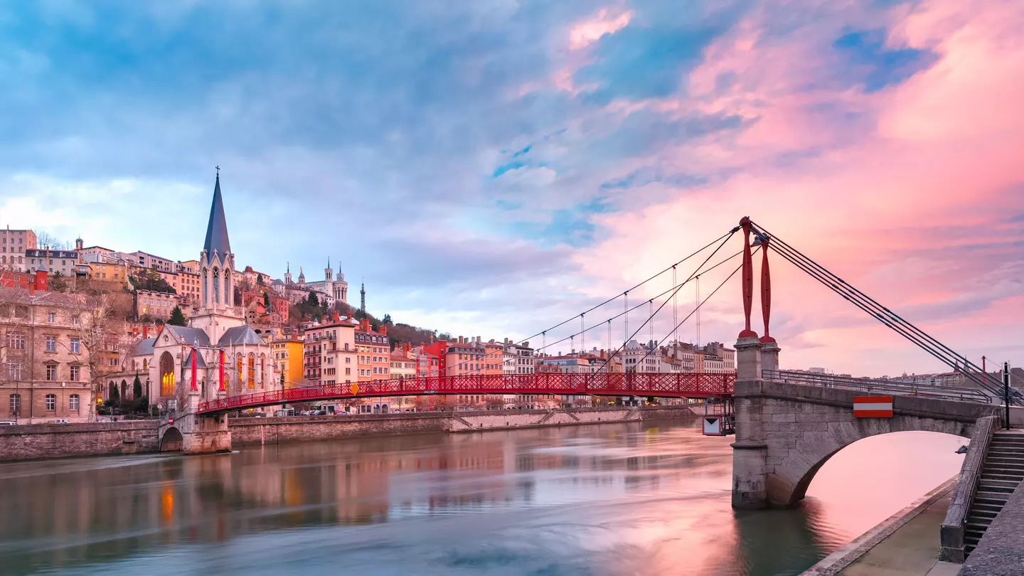 Saint Georges church and a red footbridge across the Saone river in Vieux-Lyon, bright pink clouds on the right in an otherwise blue sky.