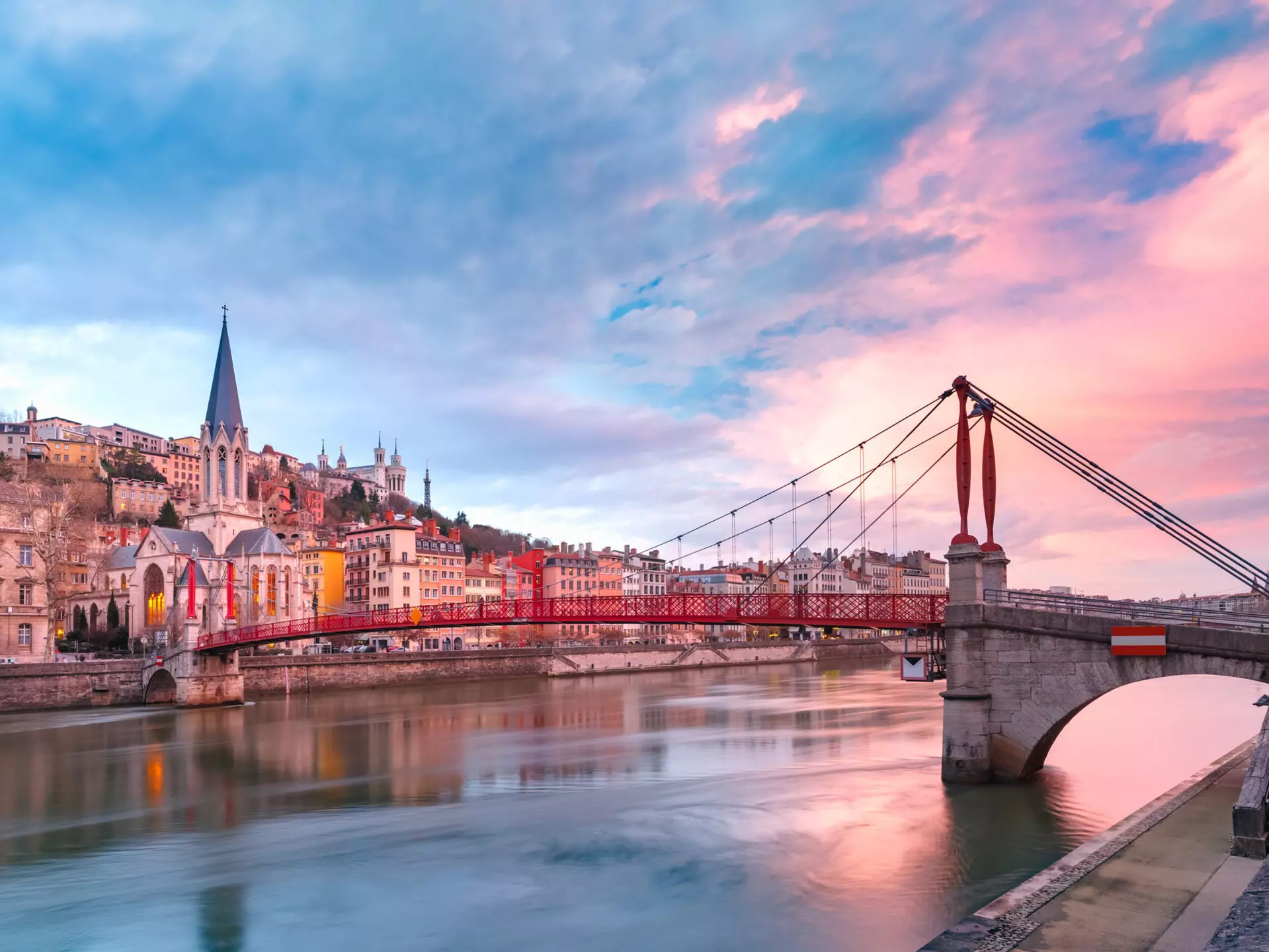 Saint Georges church and a red footbridge across the Saone river in Vieux-Lyon, bright pink clouds on the right in an otherwise blue sky.