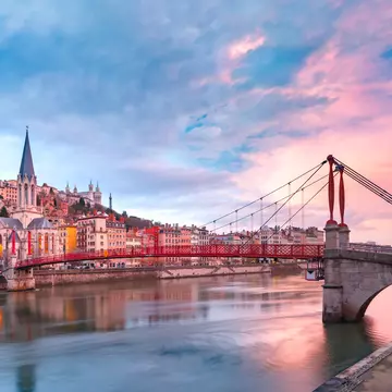 Saint Georges church and a red footbridge across the Saone river in Vieux-Lyon, bright pink clouds on the right in an otherwise blue sky.