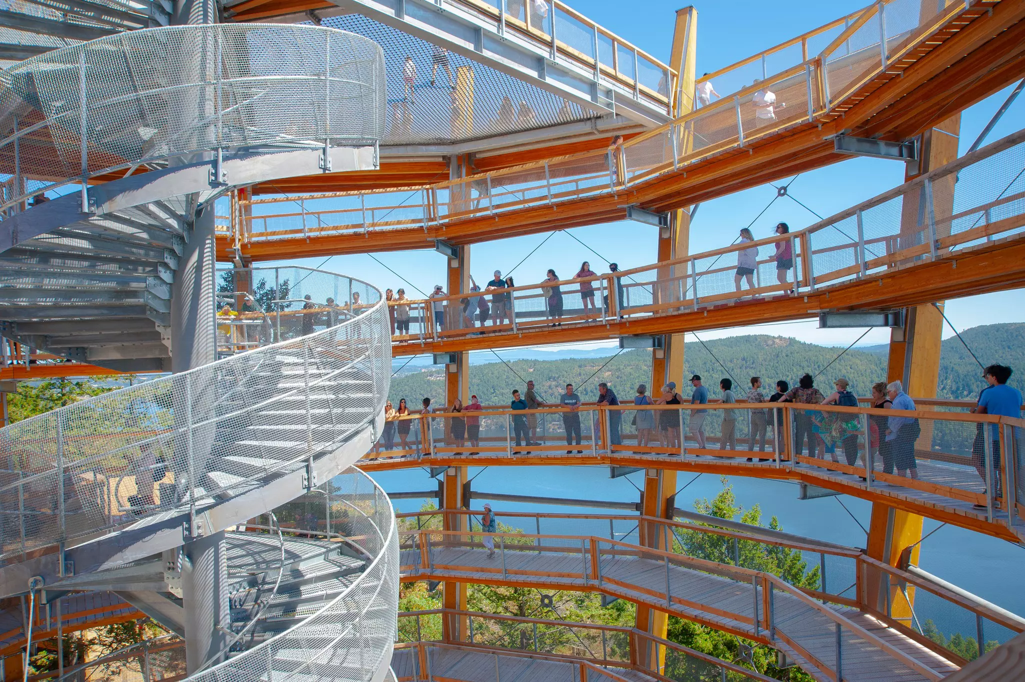 A round open-sided walkway spirals around a central staircase with views of a waterway and forest-covered islands.