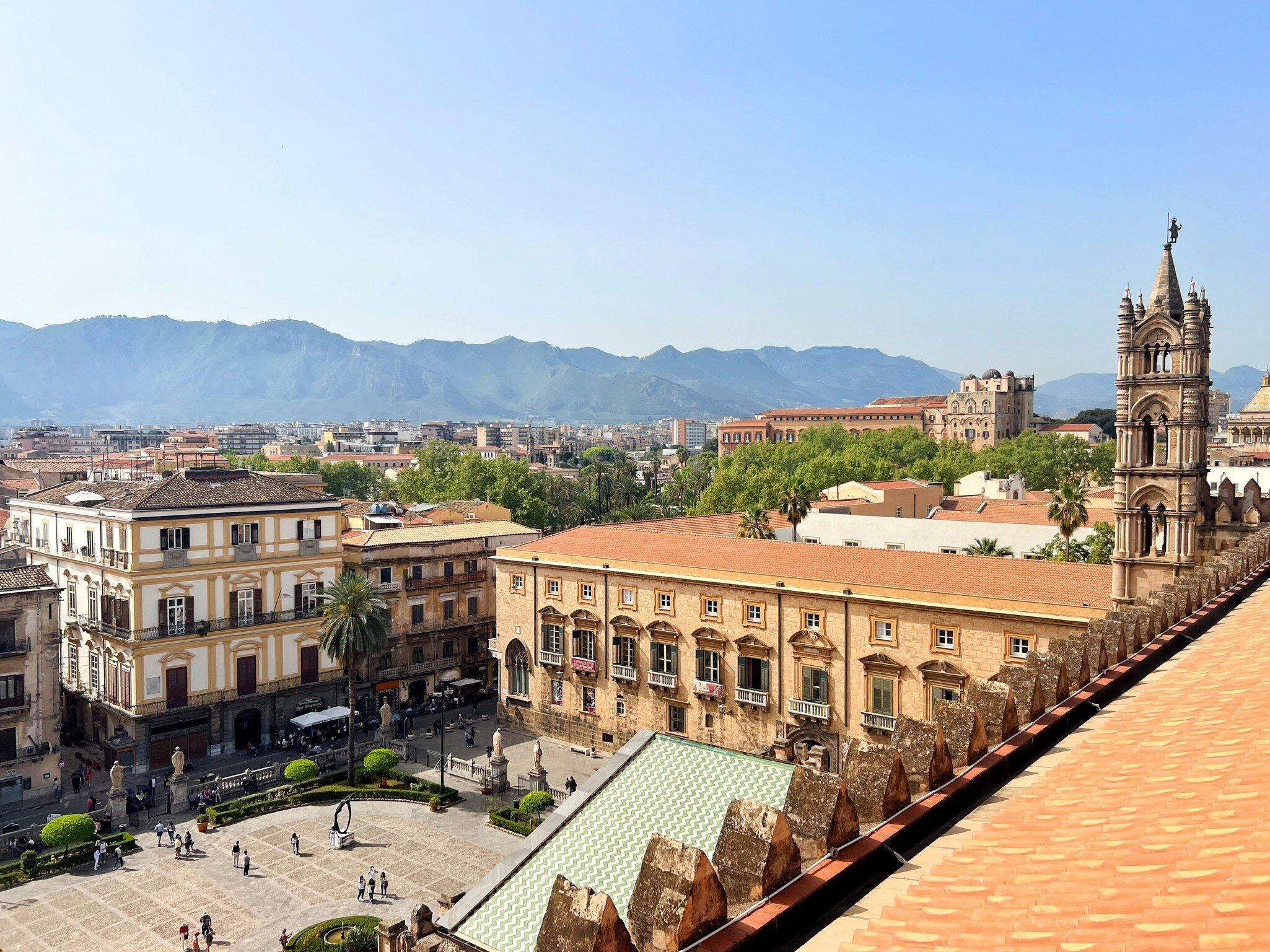Rooftop view of a long honey-coloured palazzo, a cathedral bell tower and palm-lined square below mountains