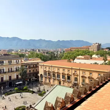 Rooftop view of a long honey-coloured palazzo, a cathedral bell tower and palm-lined square below mountains