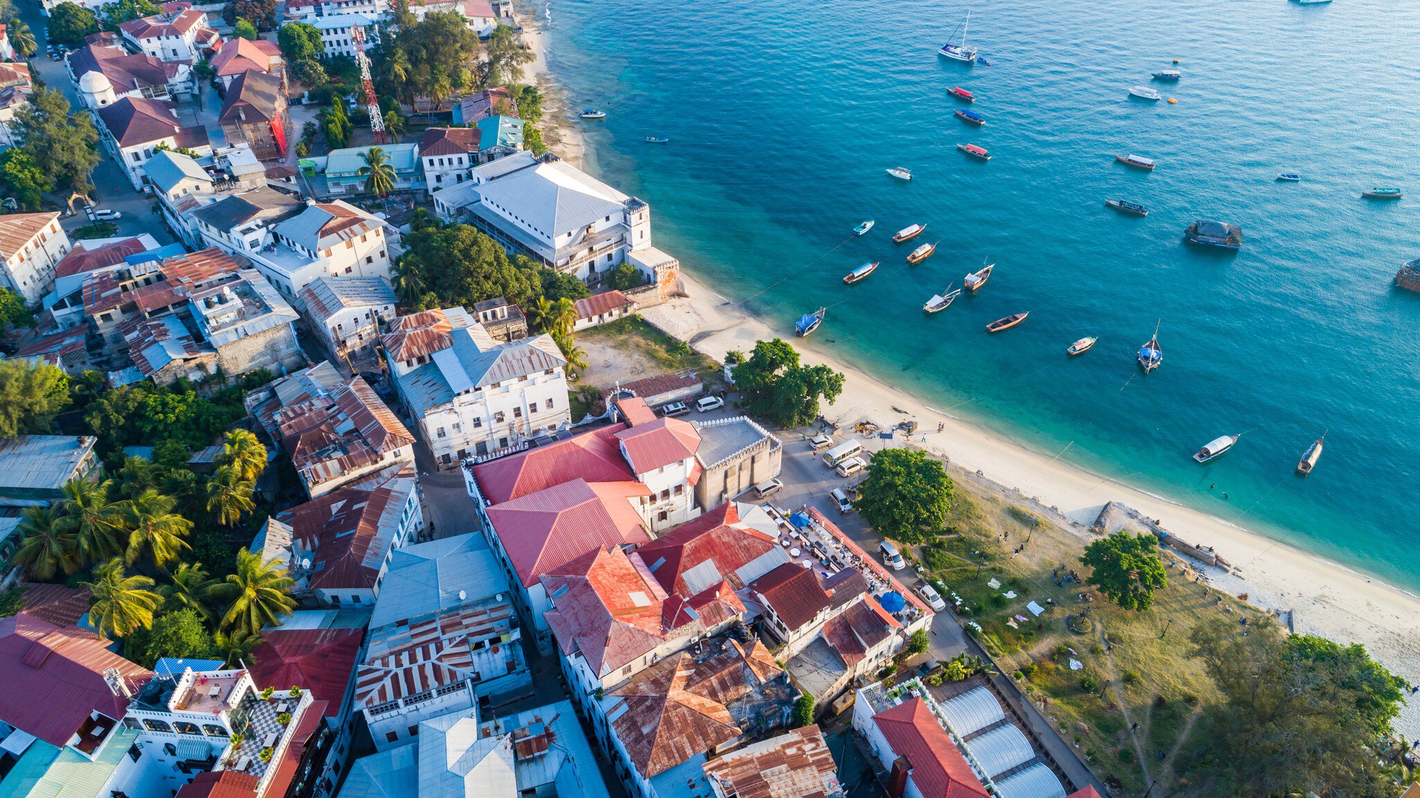 Red-roofed buildings line a sandy beach with many small boats moored in the blue ocean on a sunny day.