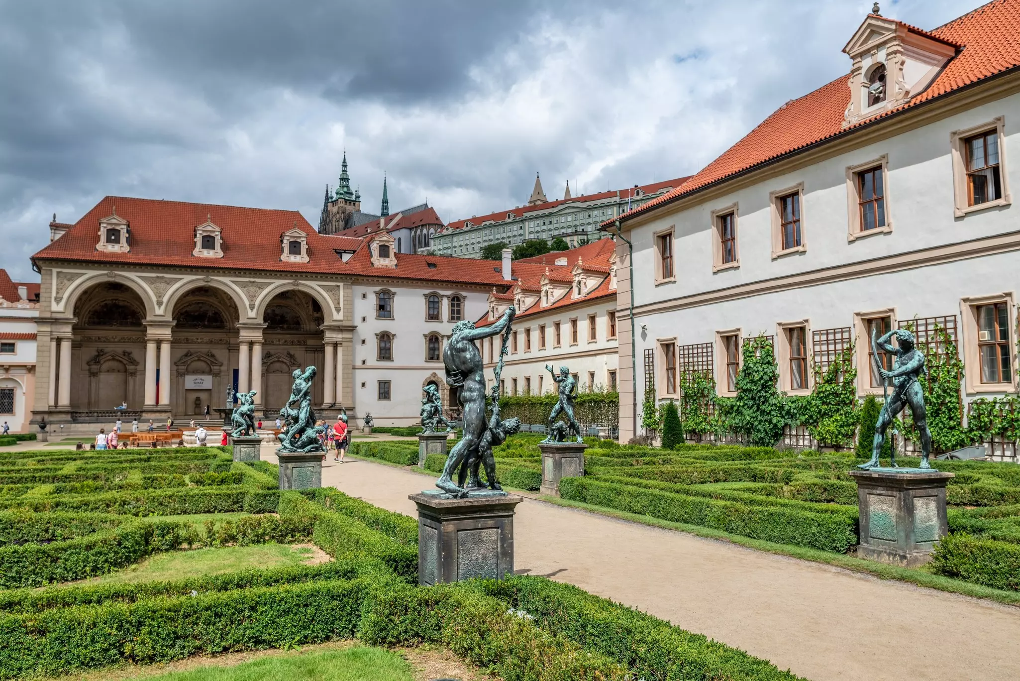 A path bordered by low hedge mazes and statues leads to a tan building with three arches.
