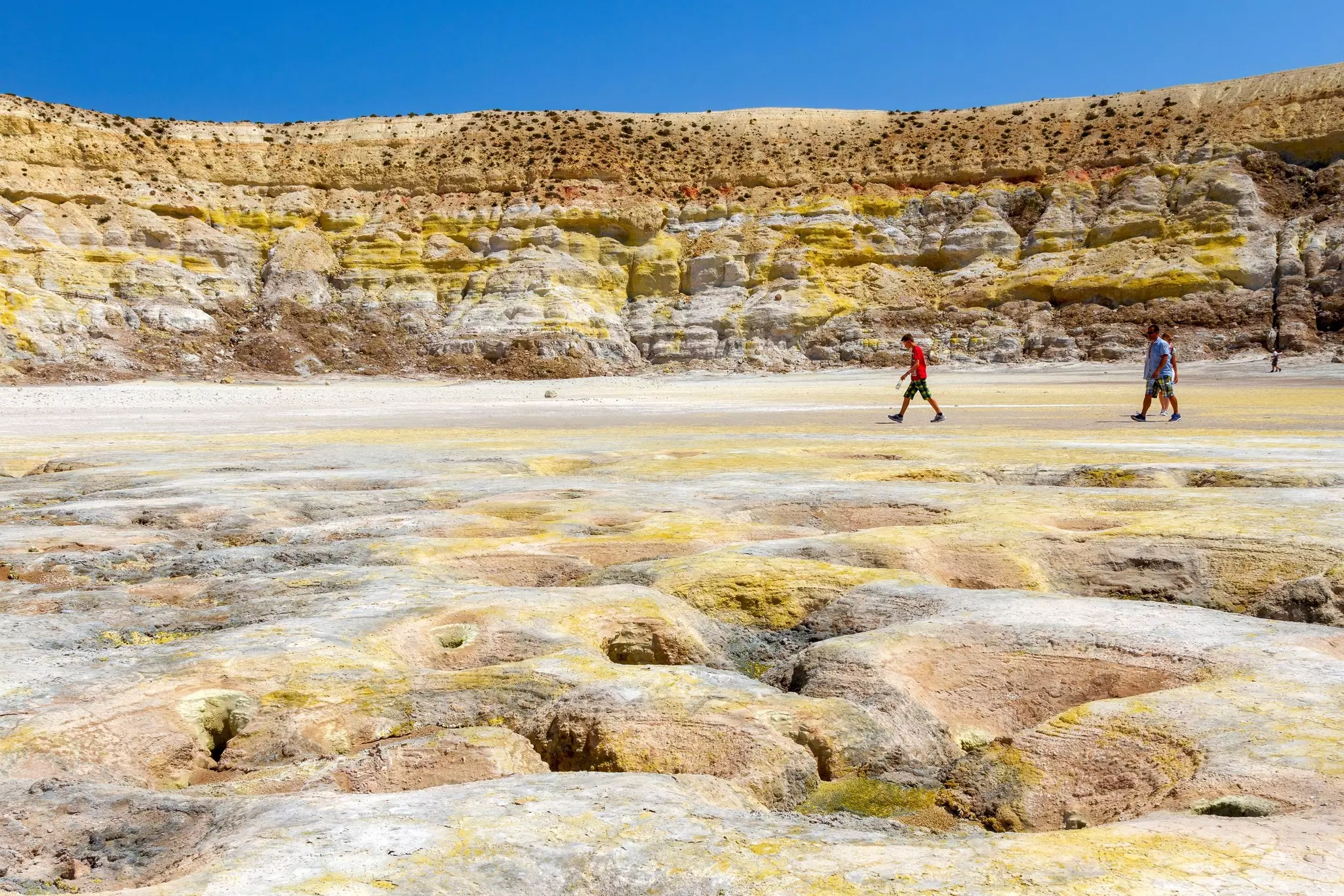 Three hikers stroll through a lunar-like landscape within a grey-yellow crater.