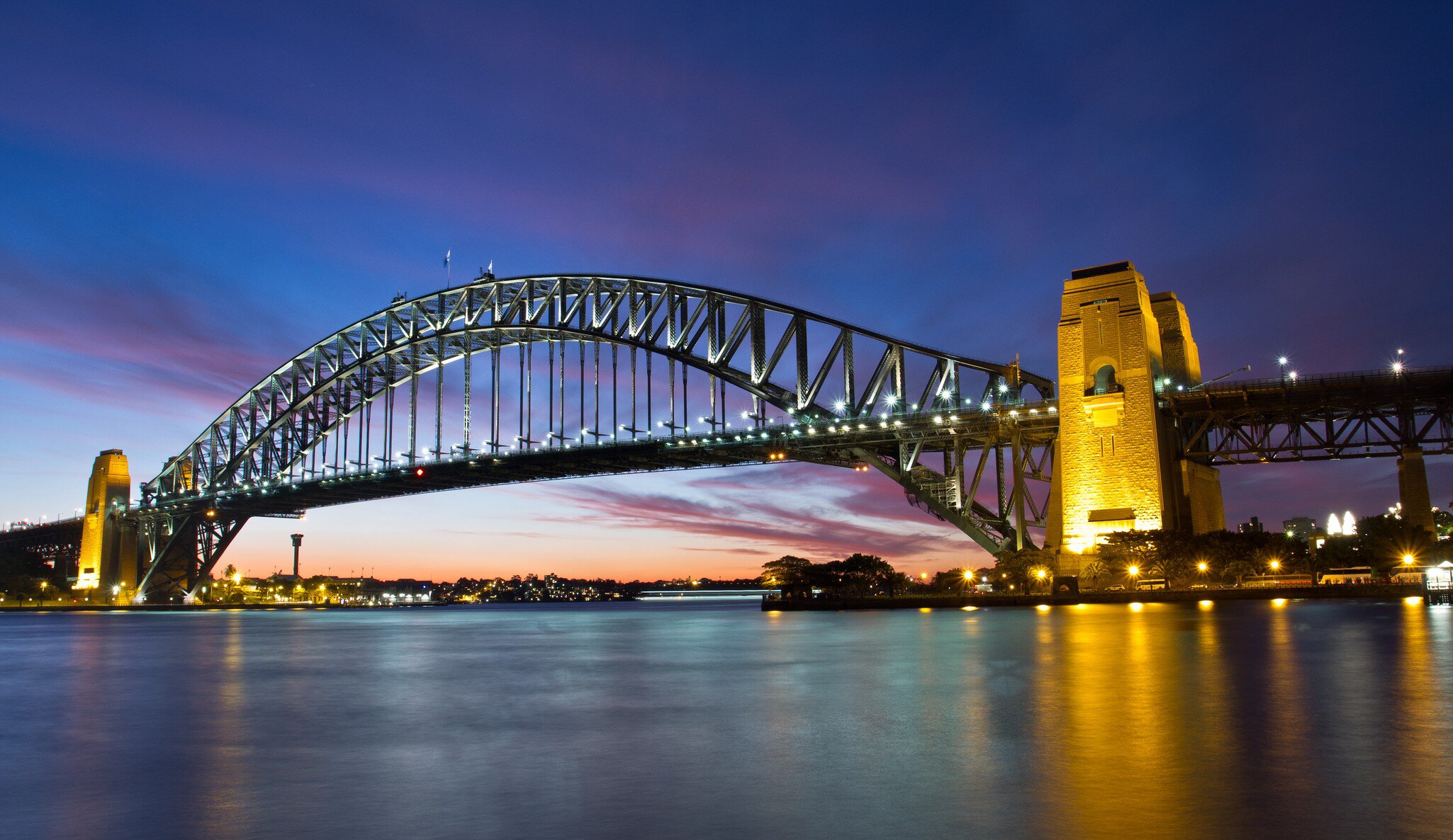 A lit steel arch bridge spanning calm water at dusk, lights reflecting below.