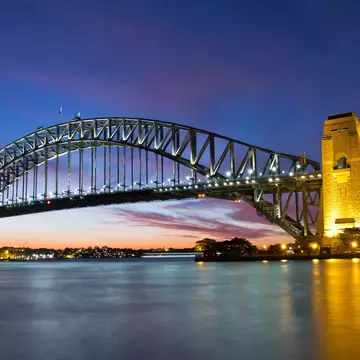 A lit steel arch bridge spanning calm water at dusk, lights reflecting below.