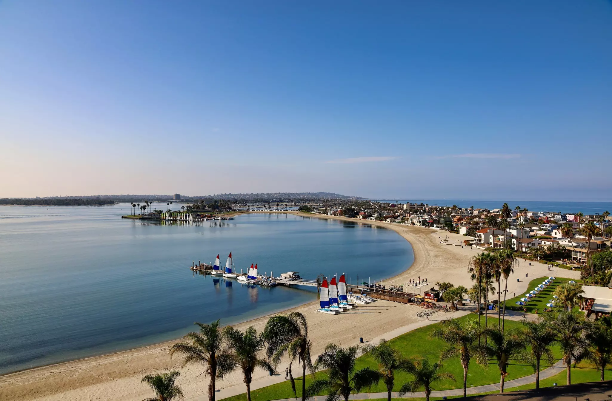 Aerial view of curved, sandy coastline with a pier and sailboats along it and on the sand, with houses to the right and the sea in the distance on a sunny day.