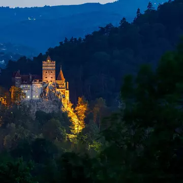 A view of a castle on a cliff top illuminated by night among shadowy hillsides