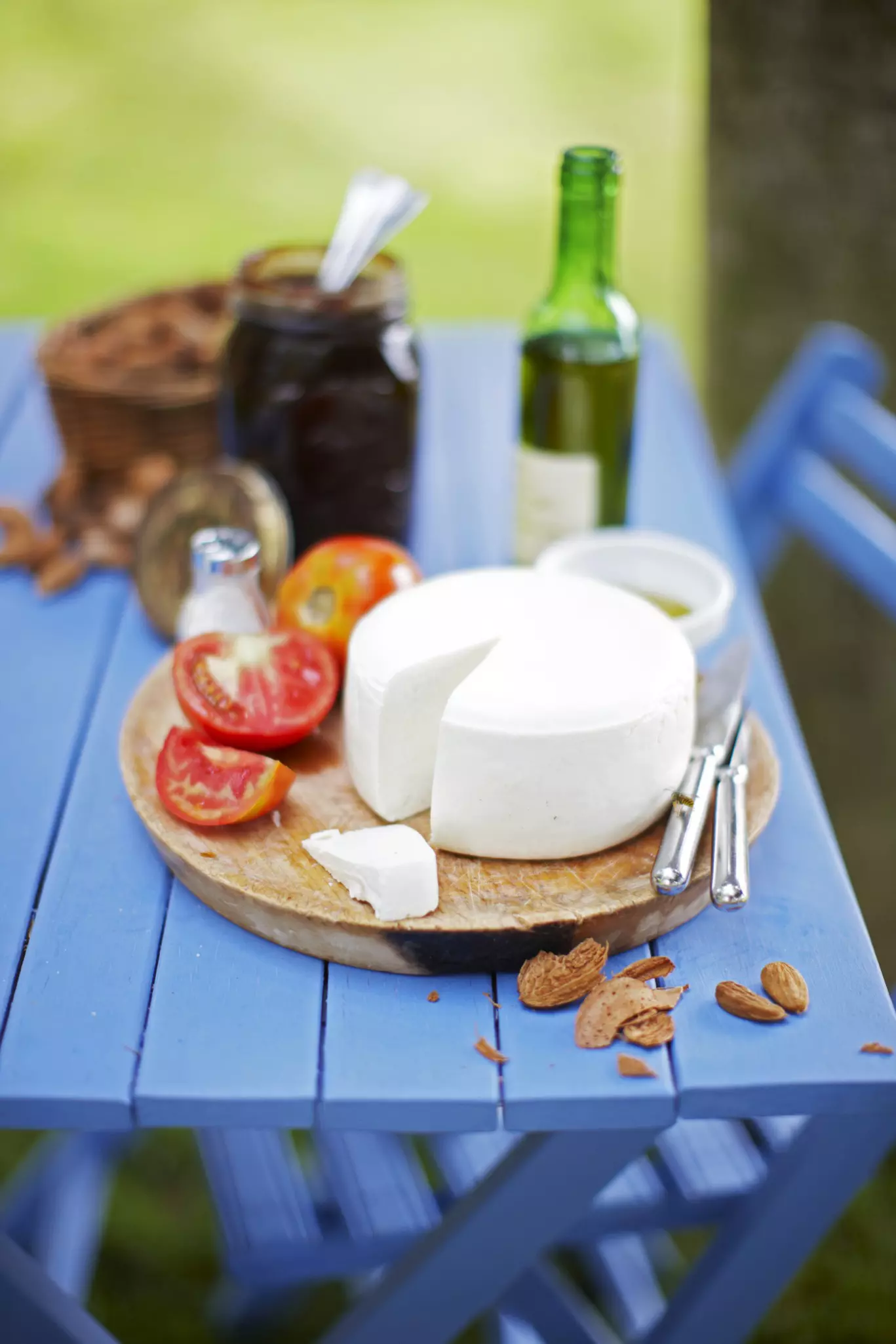 A wheel of goat cheese and tomatoes are displayed alongside a bottle of wine on a blue table.