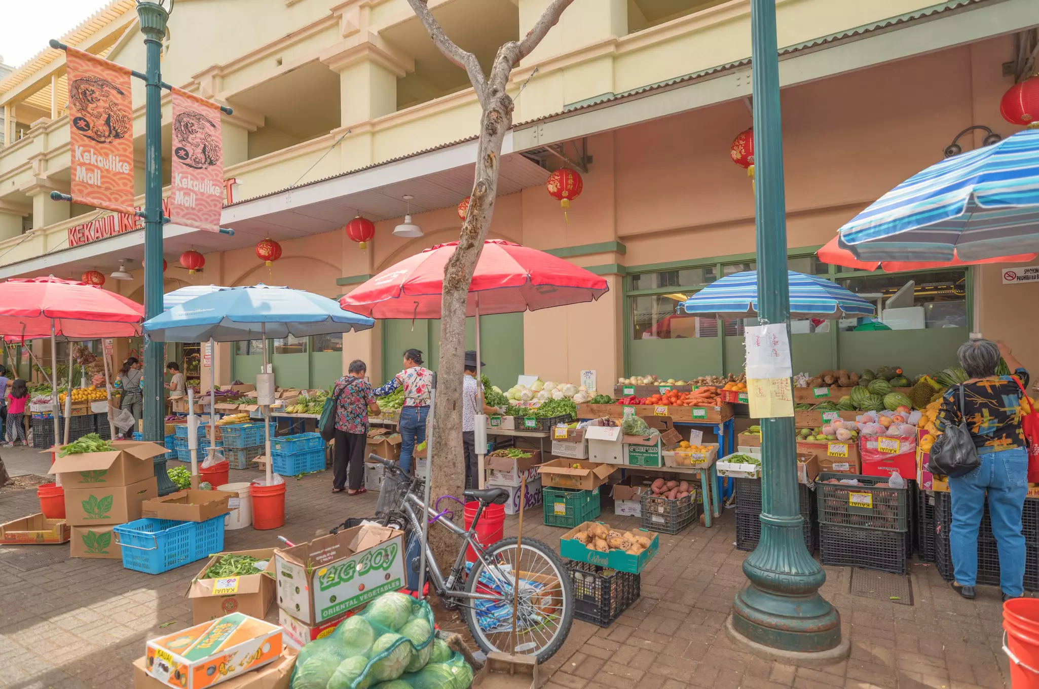 Produce tables and crates lined up under colorful umbrellas outside a building. Two banners on a lampost above them read "Kekaulike Mall."