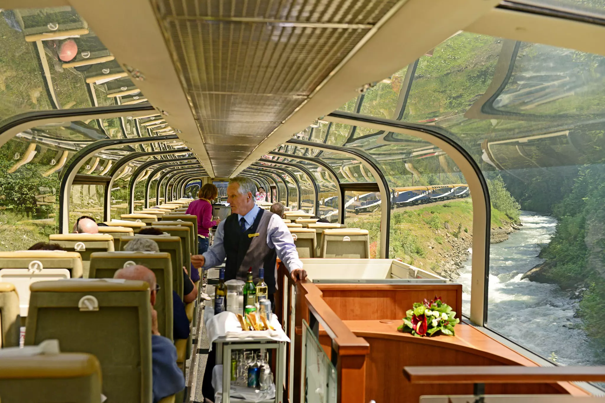 An attendant wheels a cart with drinks down the aisle of a train observation car. Passengers sit at seats overlooking a river and green mountains.