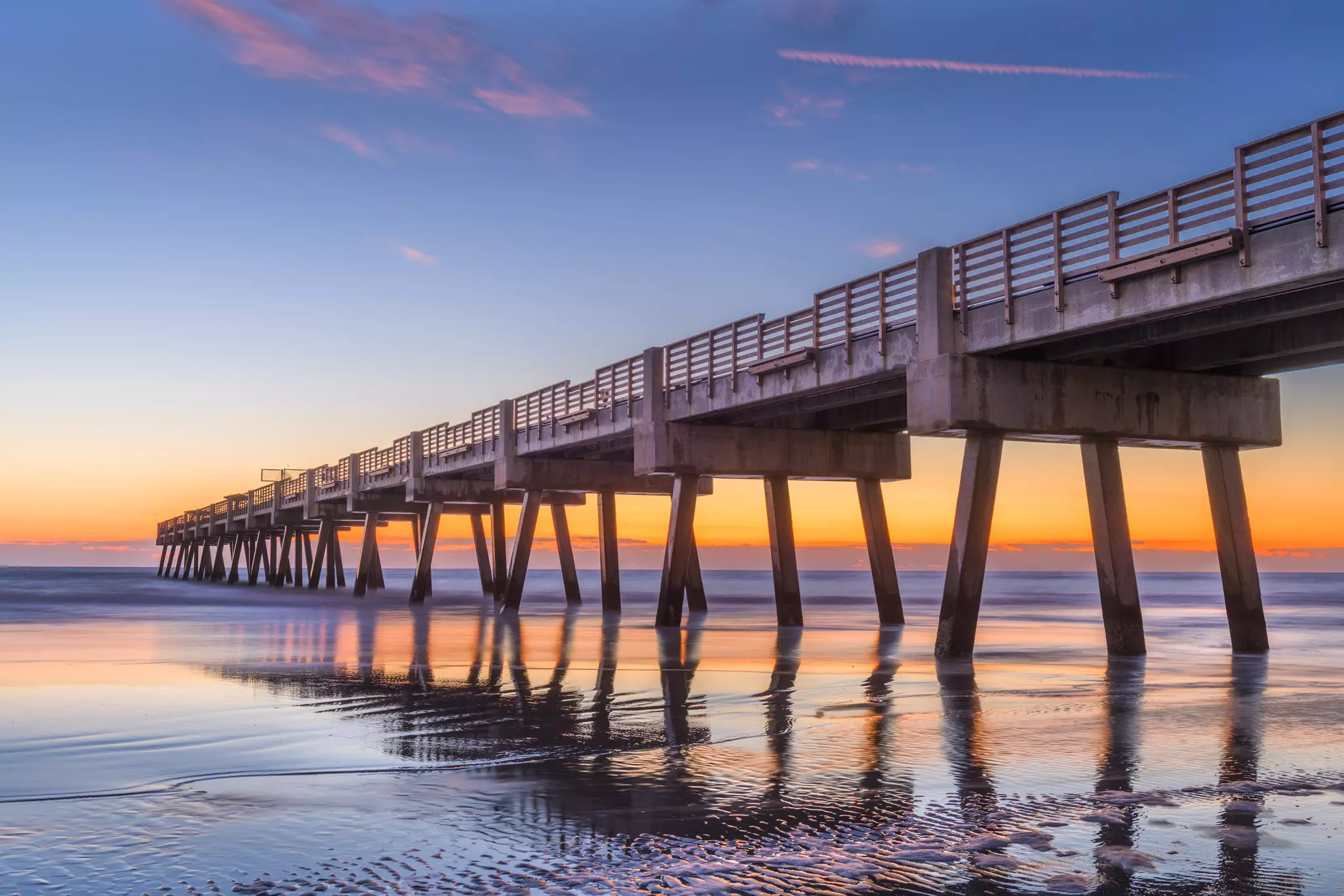 Bands of color are seen on the horizon at dawn by the ocean. A long pier on pilings sticks out into the water.