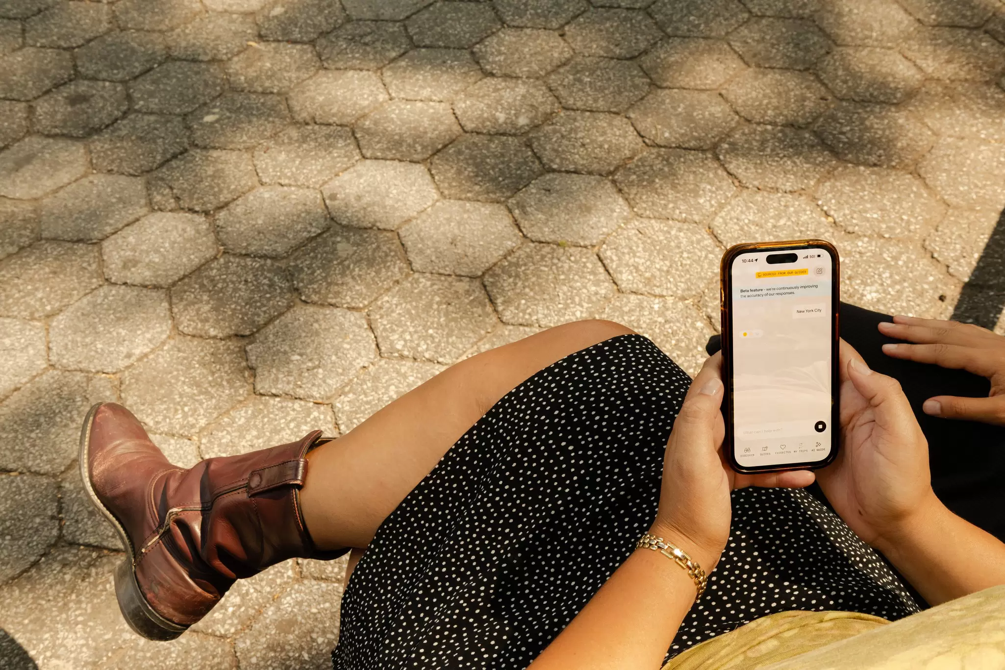 Shot from behind of a woman looking at her phone while seated in a square in NYC with dappled sunlight on the paving stones