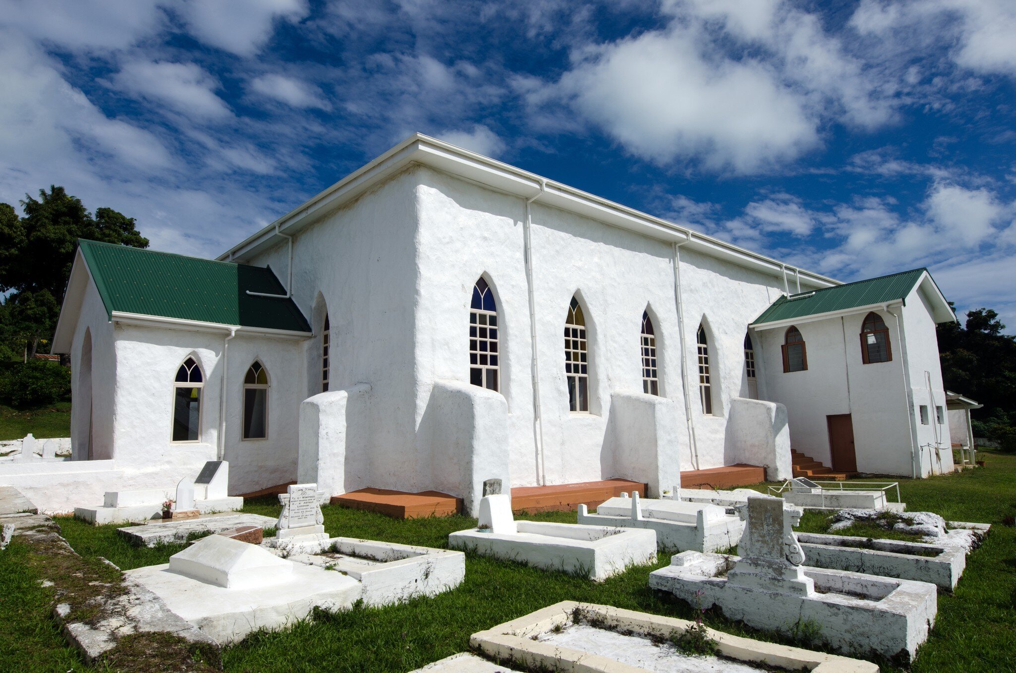 A whitewashed church with a green tin roof and pointed windows, standing among whitewashed tombs on cut grass.