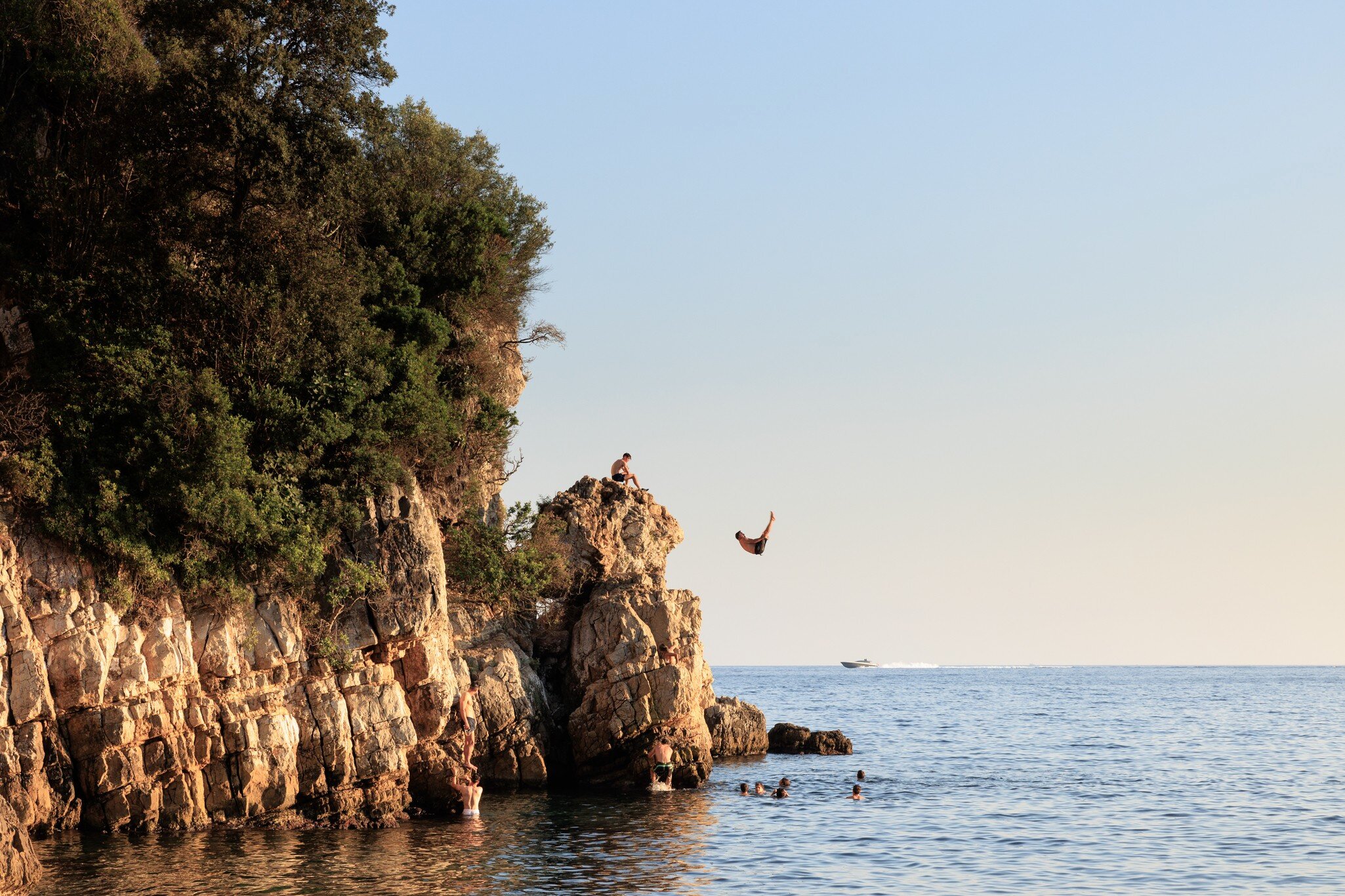 Swimmer leaps from a rocky, tree-topped headland into calm sea near Antibes, France, as others tread water below