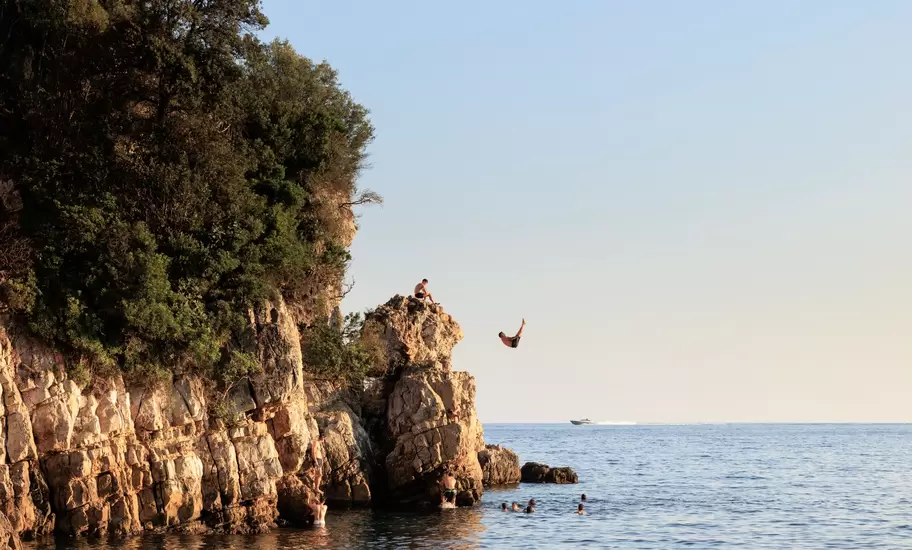 Swimmer leaps from a rocky, tree-topped headland into calm sea near Antibes, France, as others tread water below