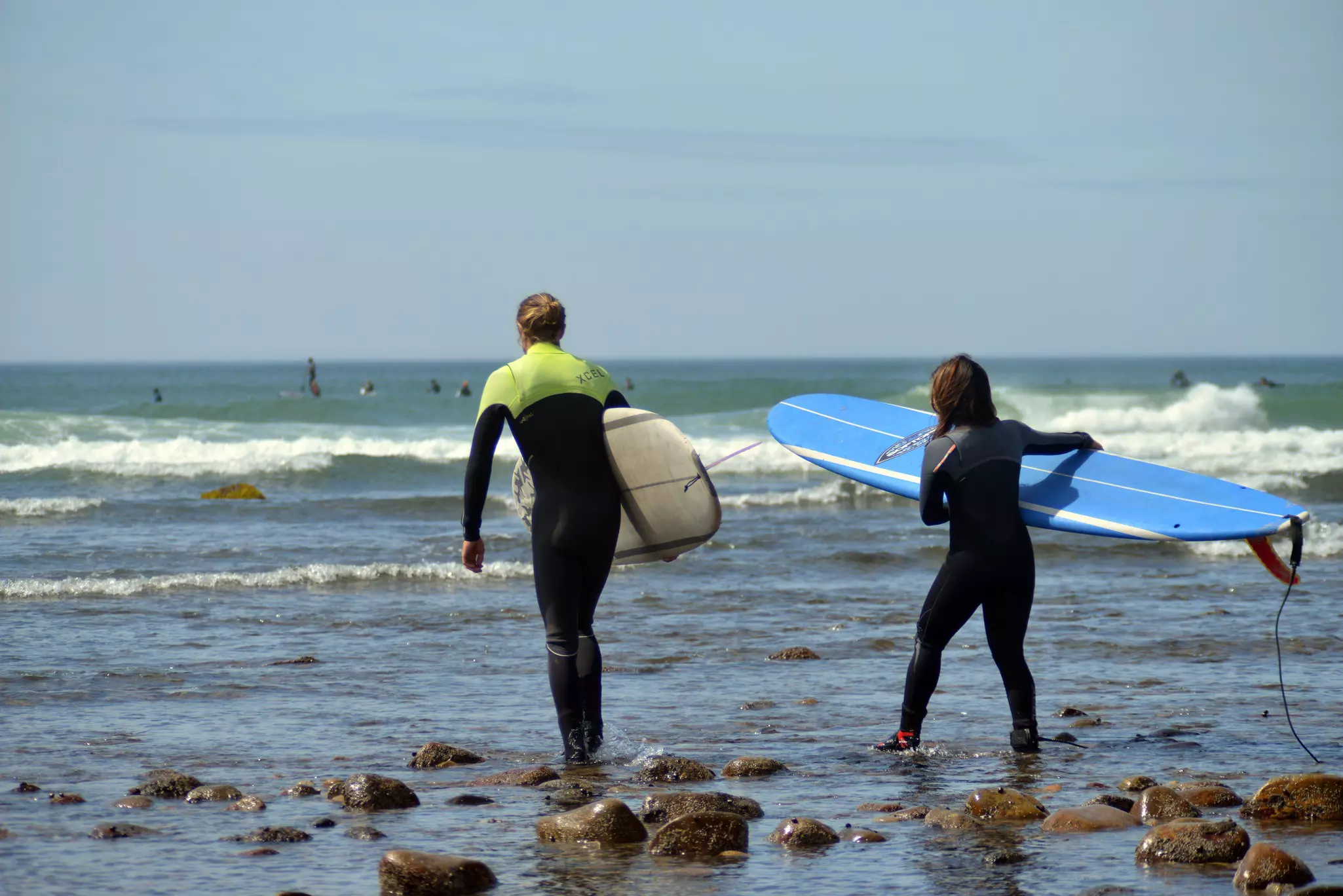Two people in wetsuits holding surfboards walking into water.