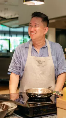 A smiling man wearing an apron stands in front of a countertop stove with a stainless pan.
