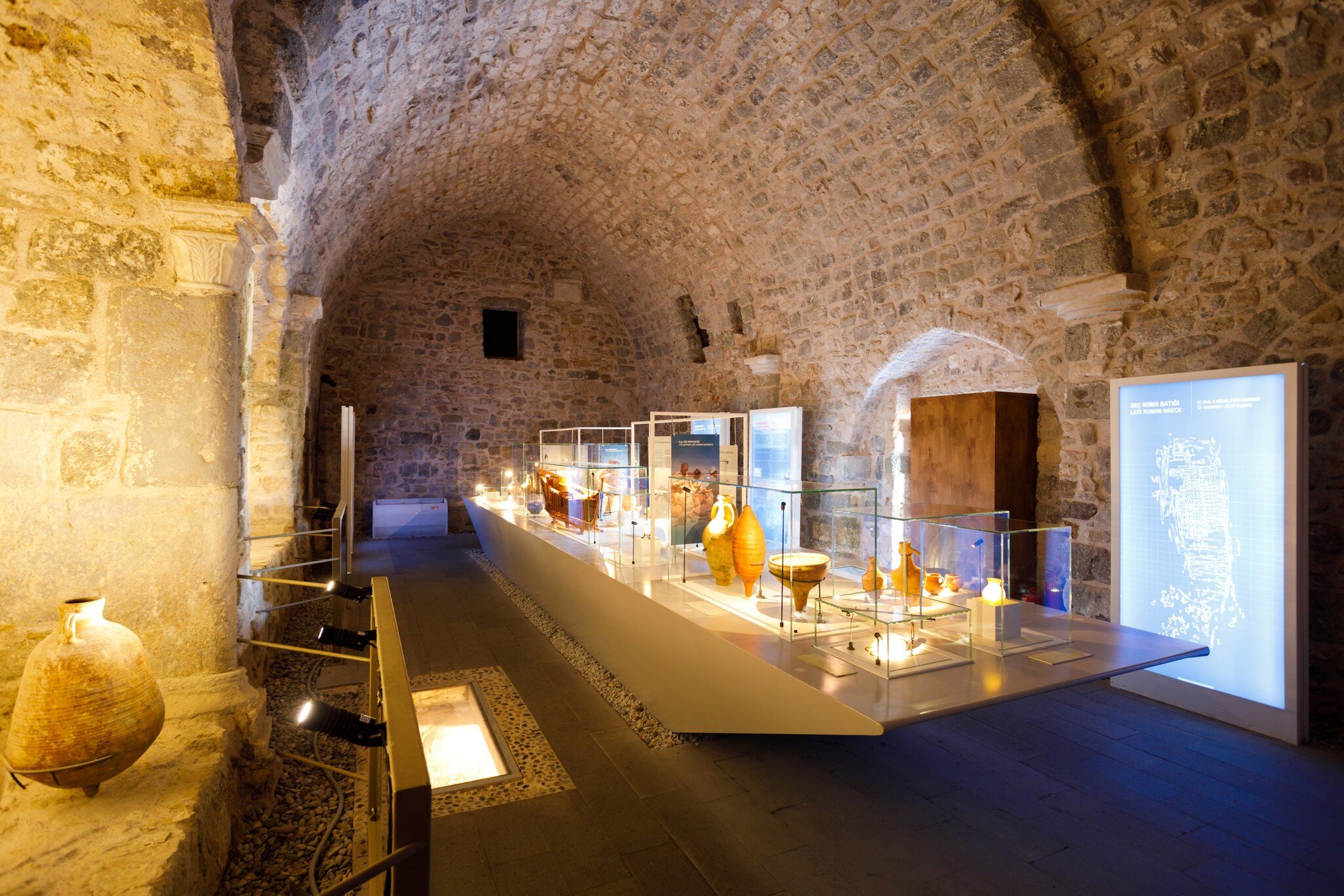 Amphorae and pottery in lit glass cases along a long display table under a rough stone barrel vault