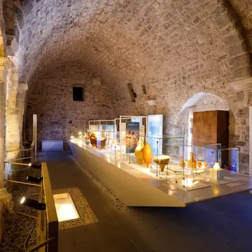 Amphorae and pottery in lit glass cases along a long display table under a rough stone barrel vault