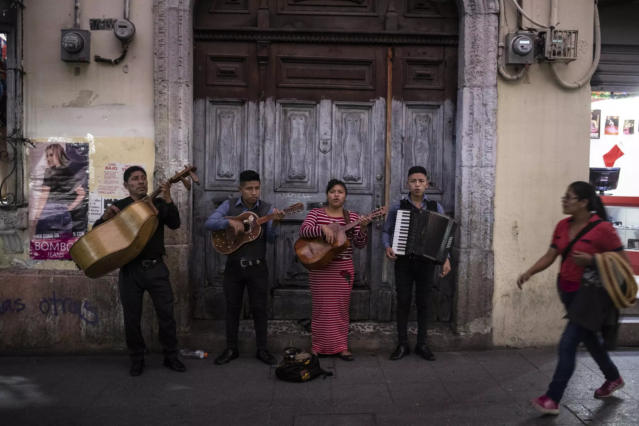 Mariachi musicians perform in front of a wooden door on a pedestrianized street in a city.