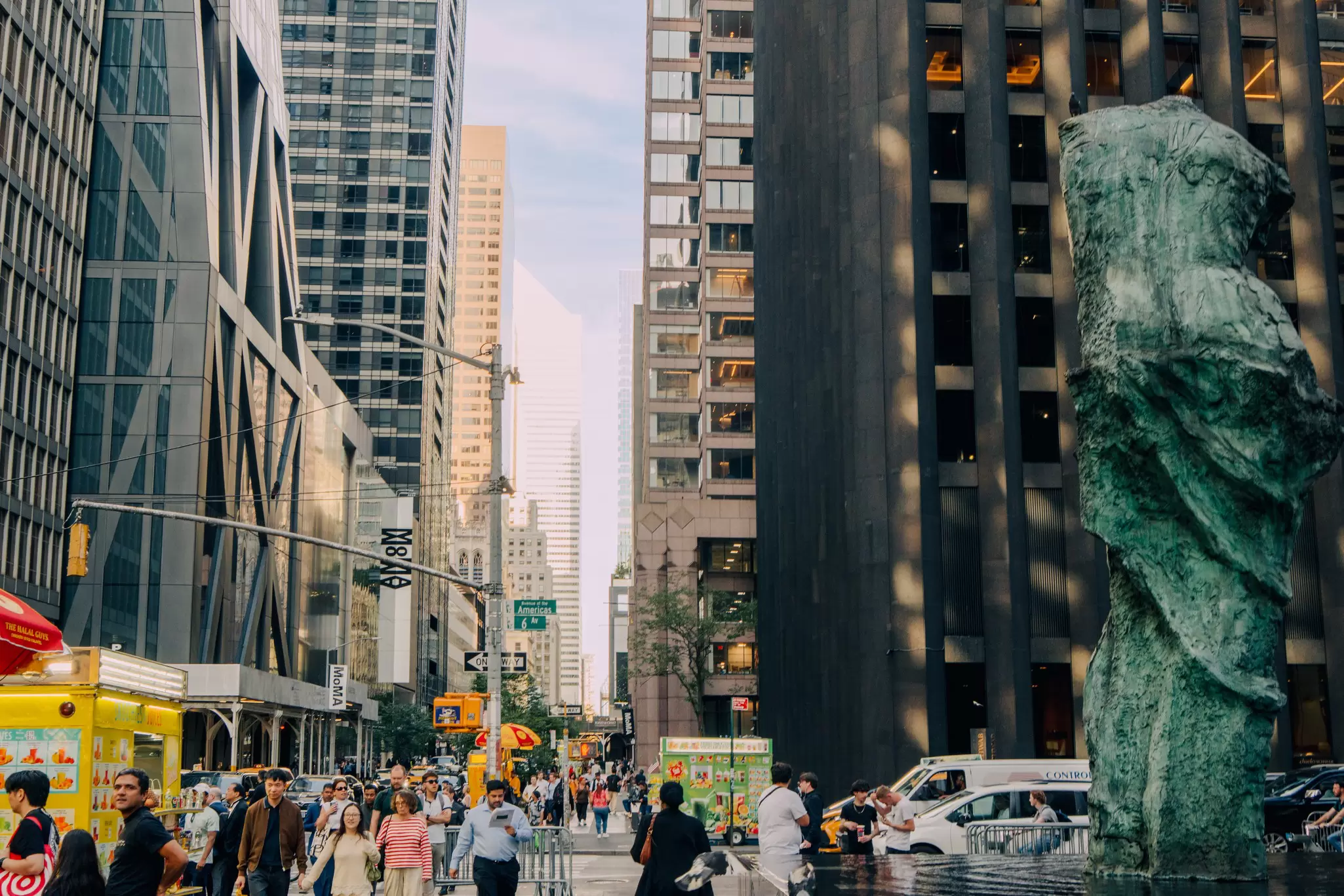 People walk past sculptures and skyscrapers on a city street