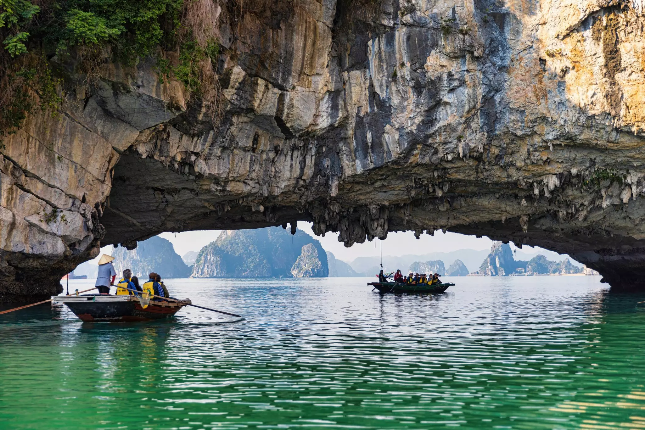Rowboats paddle under a limestone arch in a tropical bay. Other karst formations rising from the water are visible in the distance.