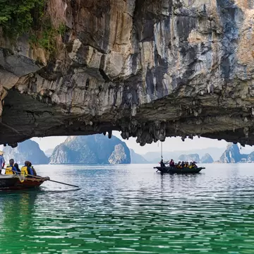 Rowboats paddle under a limestone arch in a tropical bay. Other karst formations rising from the water are visible in the distance.