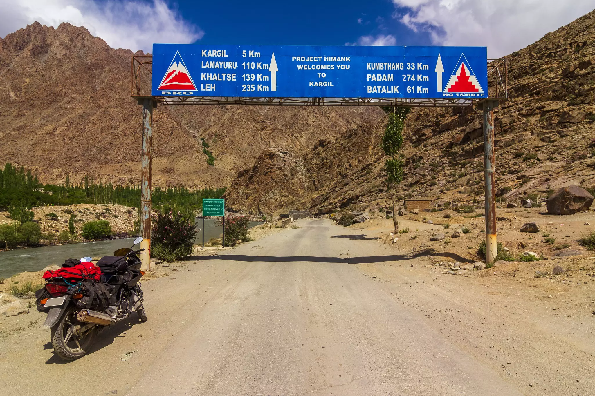A motorbike parked by a sign above a dirt road that indicates distances including 235km to Leh