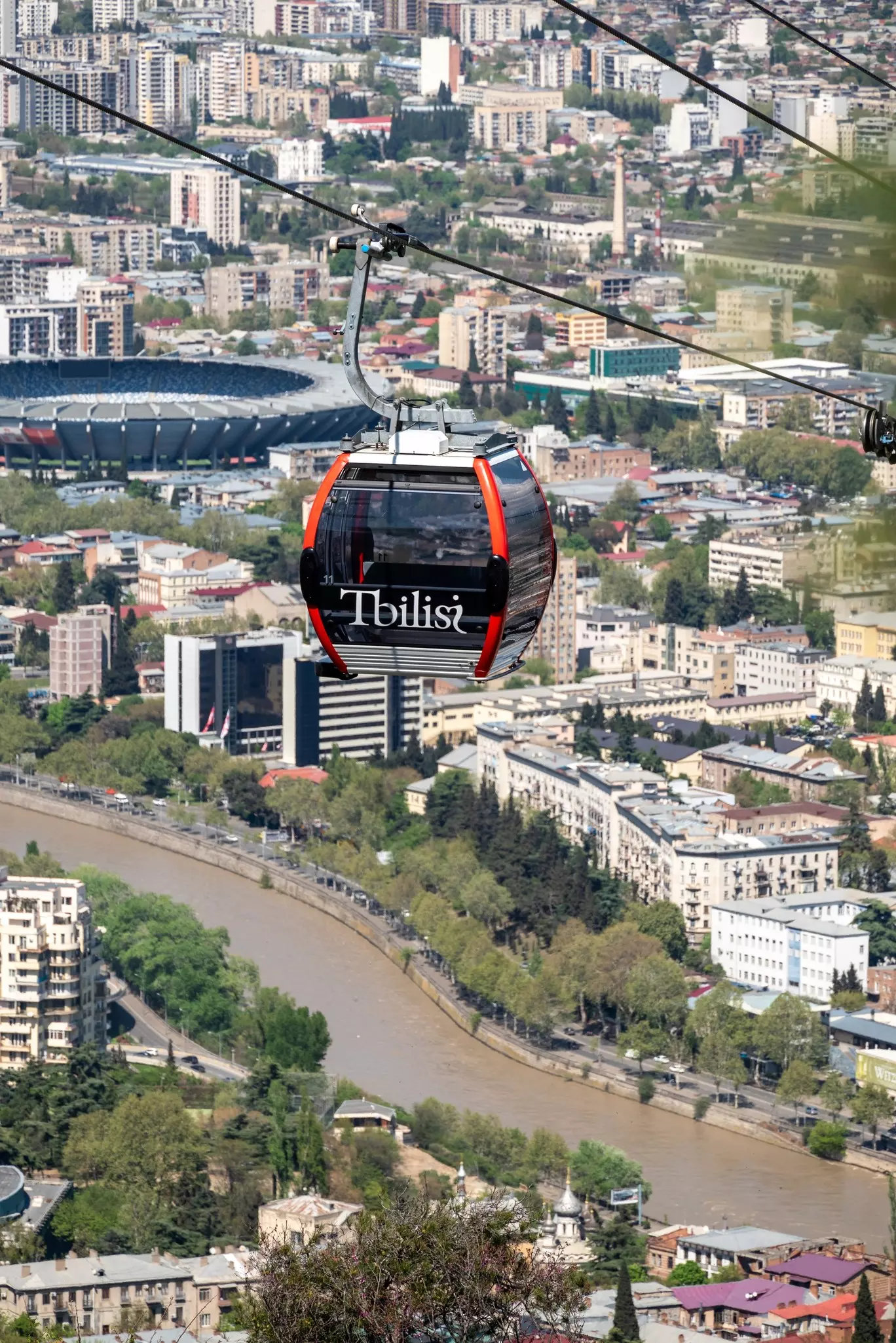 A sleek black cable car with red trim with "Tbilisi" in white letters on the side glides over the city