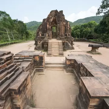 Ruined red-brick temple foundations lead to a crumbling brick tower, with green wooded hills behind.