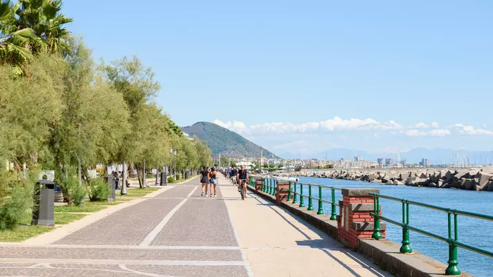 People on a long walkway by the sea; there is a mountain and a town in the distance, and a park with trees and grass on the other side of the path.