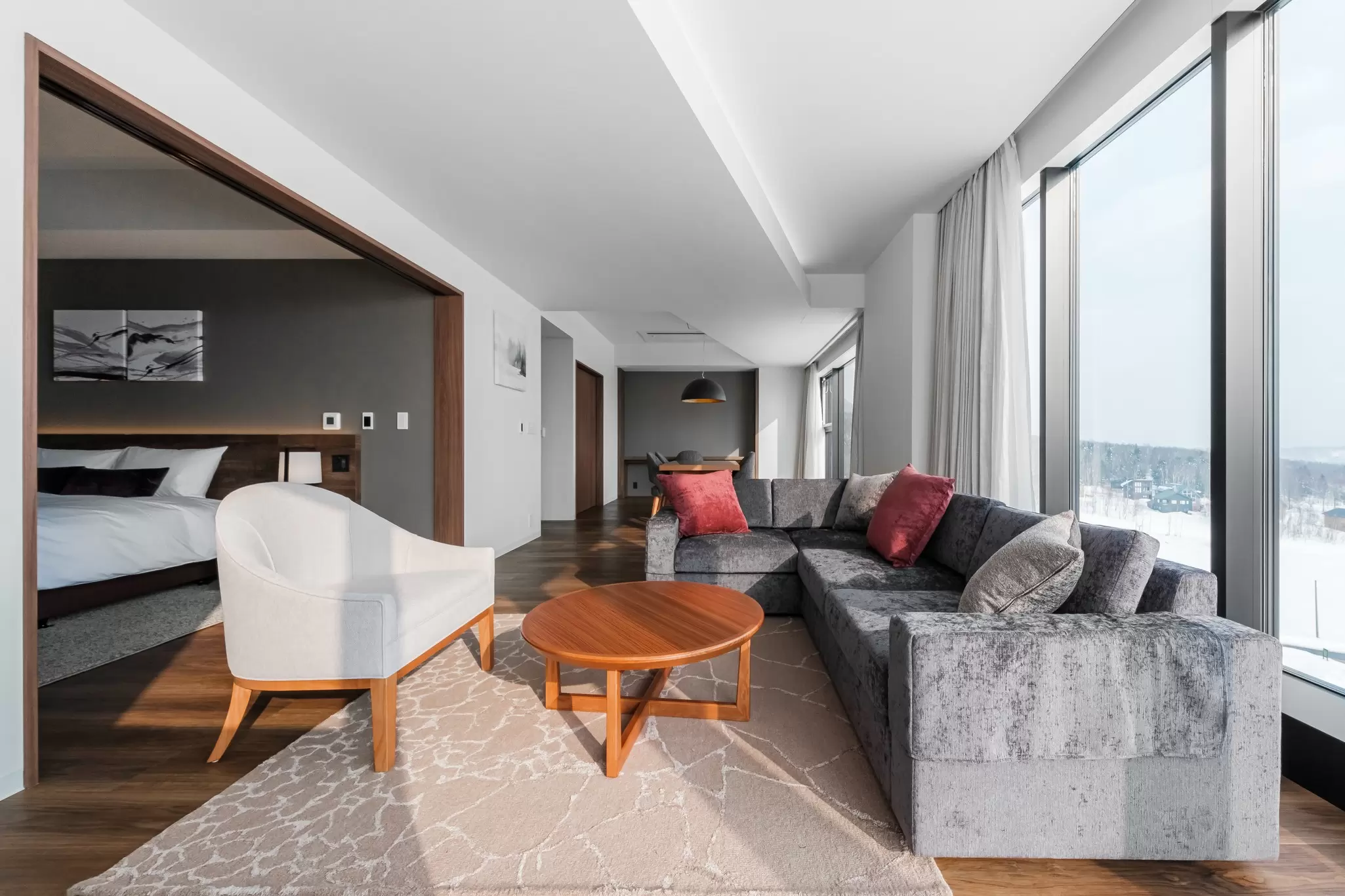 A hotel room living area with an L-shaped couch, coffee table and armchair, with the bedroom in the background, and a snowy landscape out the large window: A hotel in Hokkaidō, Japan