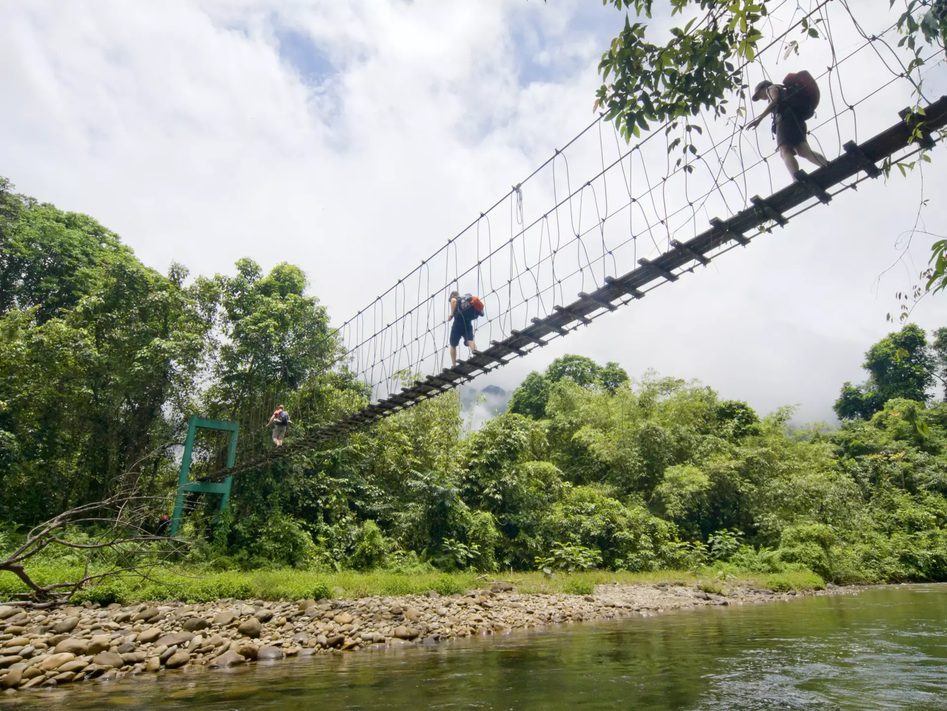 Three tourists with backpacks crossing a suspension bridge over water in Malaysia.