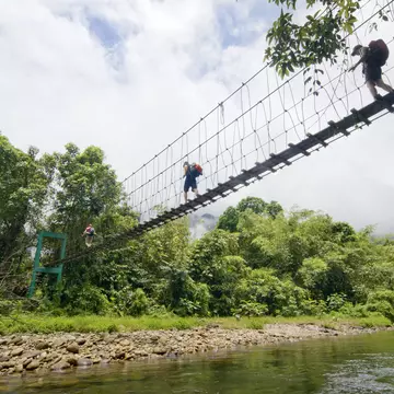 Three tourists with backpacks crossing a suspension bridge over water in Malaysia.