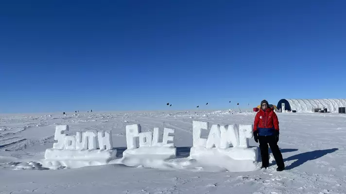 A woman stands in front of a sign that appears to be made out of snow saying "South Pole Camp"