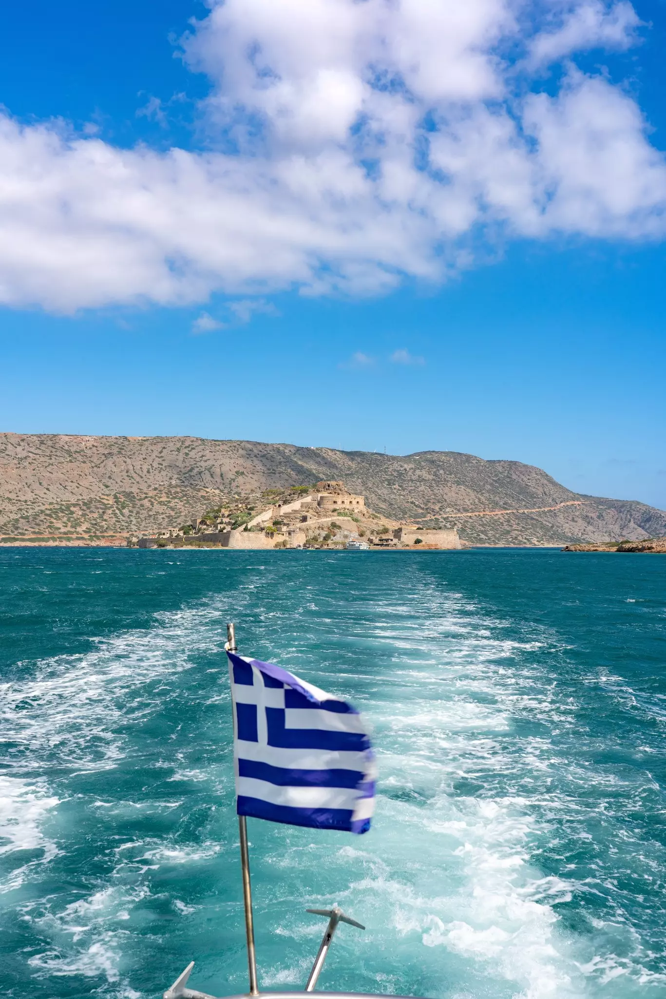 A vibrant Greek flag flutters on a boat approaching Spinalonga Island, Crete. The turquoise sea contrasts beautifully with rugged terrain and ancient ruins under a bright sky, License Type: media, Download Time: 2025-04-16T15:07:31.000Z, User: katelyn.perry_lonelyplanet, Editorial: false, purchase_order: 65050 - Digital Destinations and Articles, job: wip, client: wip, other: Katelyn Perry