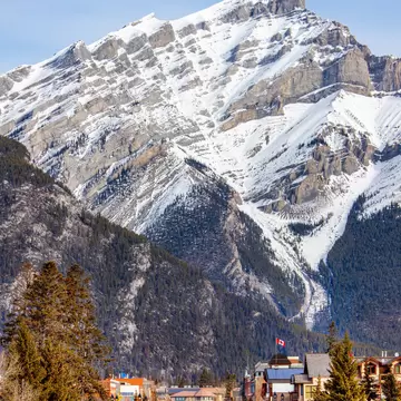 Road through the town of Banff with pine trees and a Canadian flag, below snowy Cascade Mountain in winter.