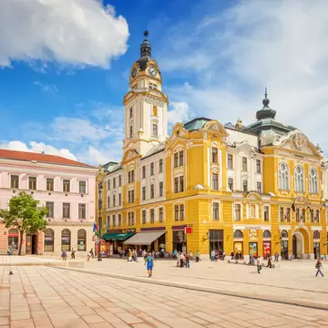 Yellow town hall with a clock tower beside a pink building and a bronze horseman statue, people crossing a paved square