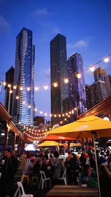 People standing and sitting at tables under yellow umbrellas in the evening; a string of white lights is overhead, and tall buildings are in the background.  