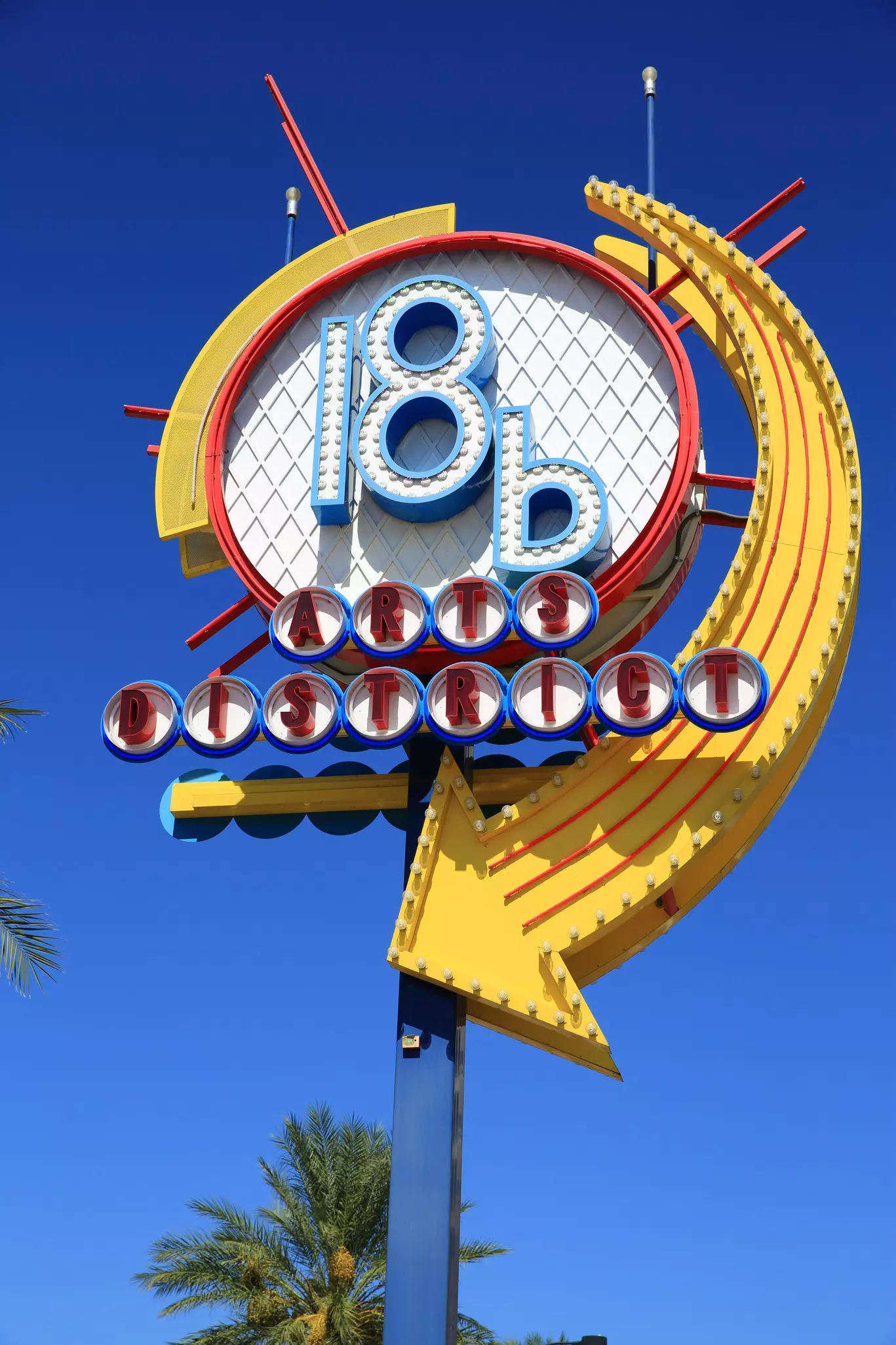 A tall sign, reminiscent of a lollypop, stands beside a palm tree on a street in Las Vegas; the sign states: 18b Arts District.