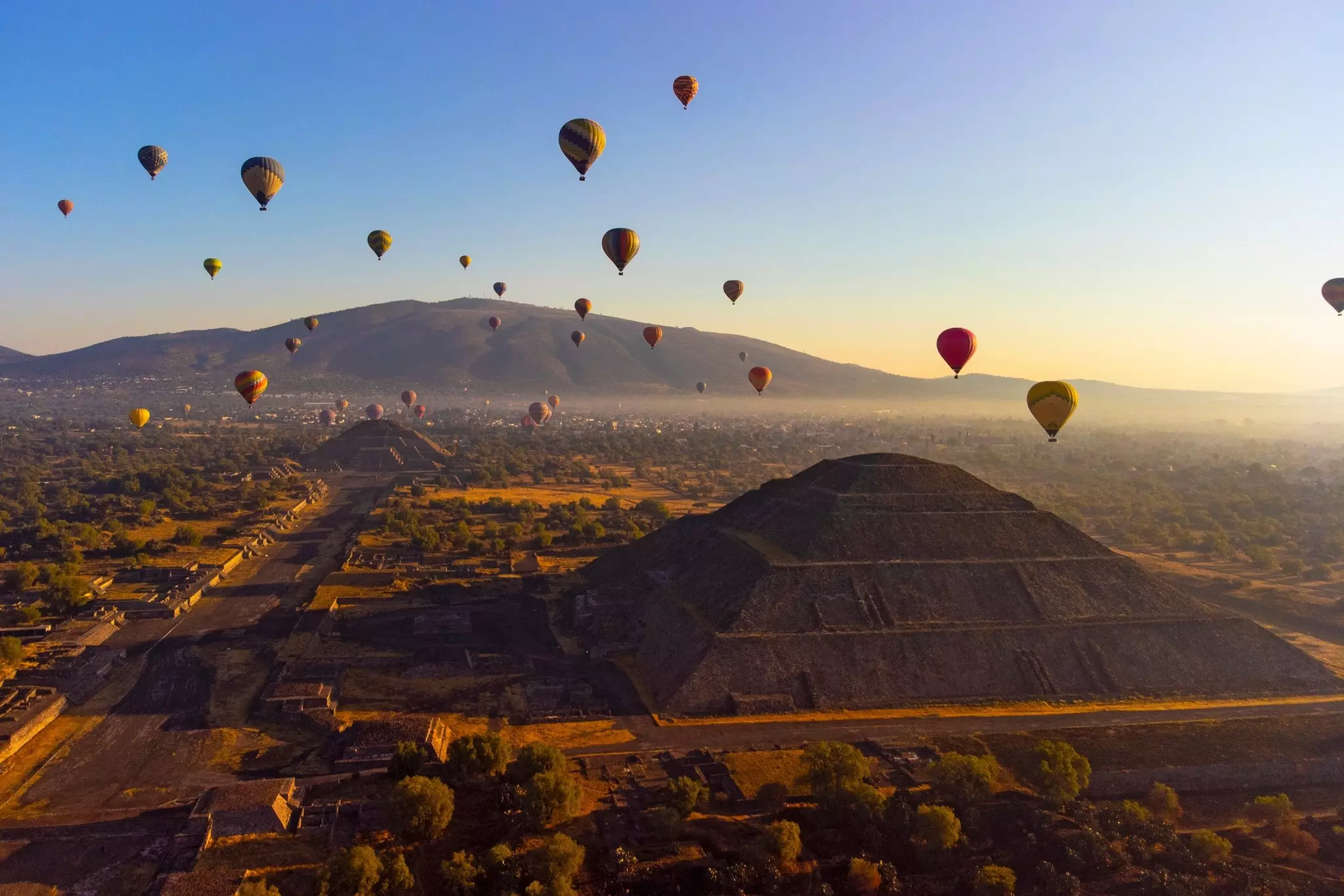 Aerial shot of hot air ballons over pyramids at sunrise with mountains in the distance.