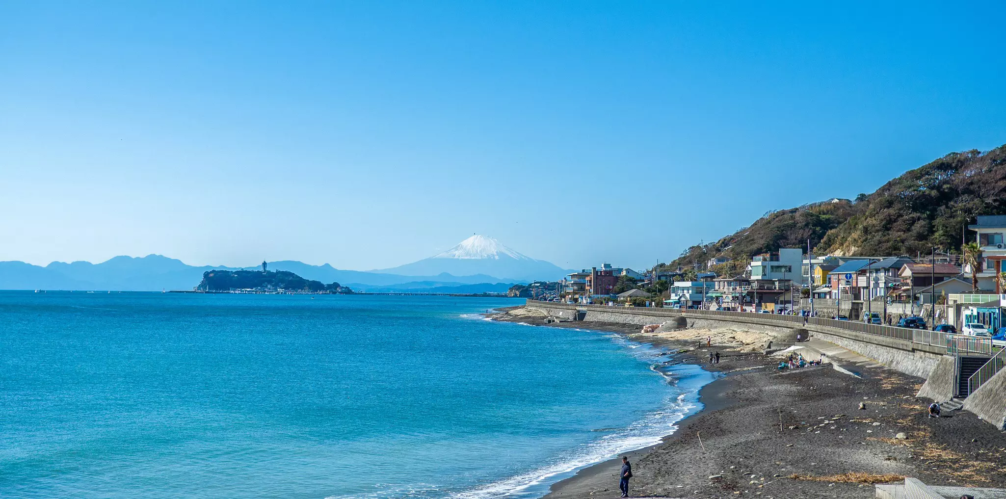 Aerial shot of curved beach bordered by a cement wall with houses beyond to the right, the ocean to the left, and a snow-capped mountain in the far distance on a sunny day.