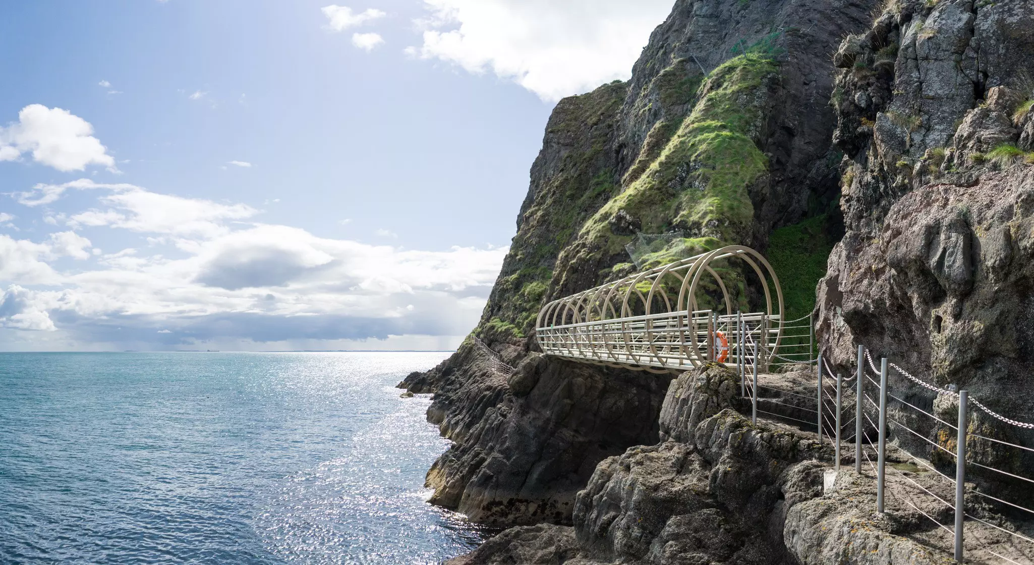 A metal walkway leads along the face of a steep cliff by the sea.