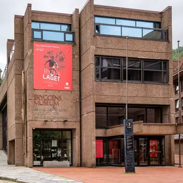A blocky pink-brown concrete museum with Bryggens Museum lettering and a red exhibition banner, under grey skies
