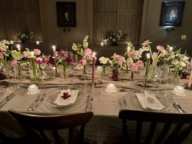A table set with clear plates that hold white napkins with a single fresh flower in the center, silverware and glasses; the center of the table has clear vases with pink flowers, purple candles and small white votives.