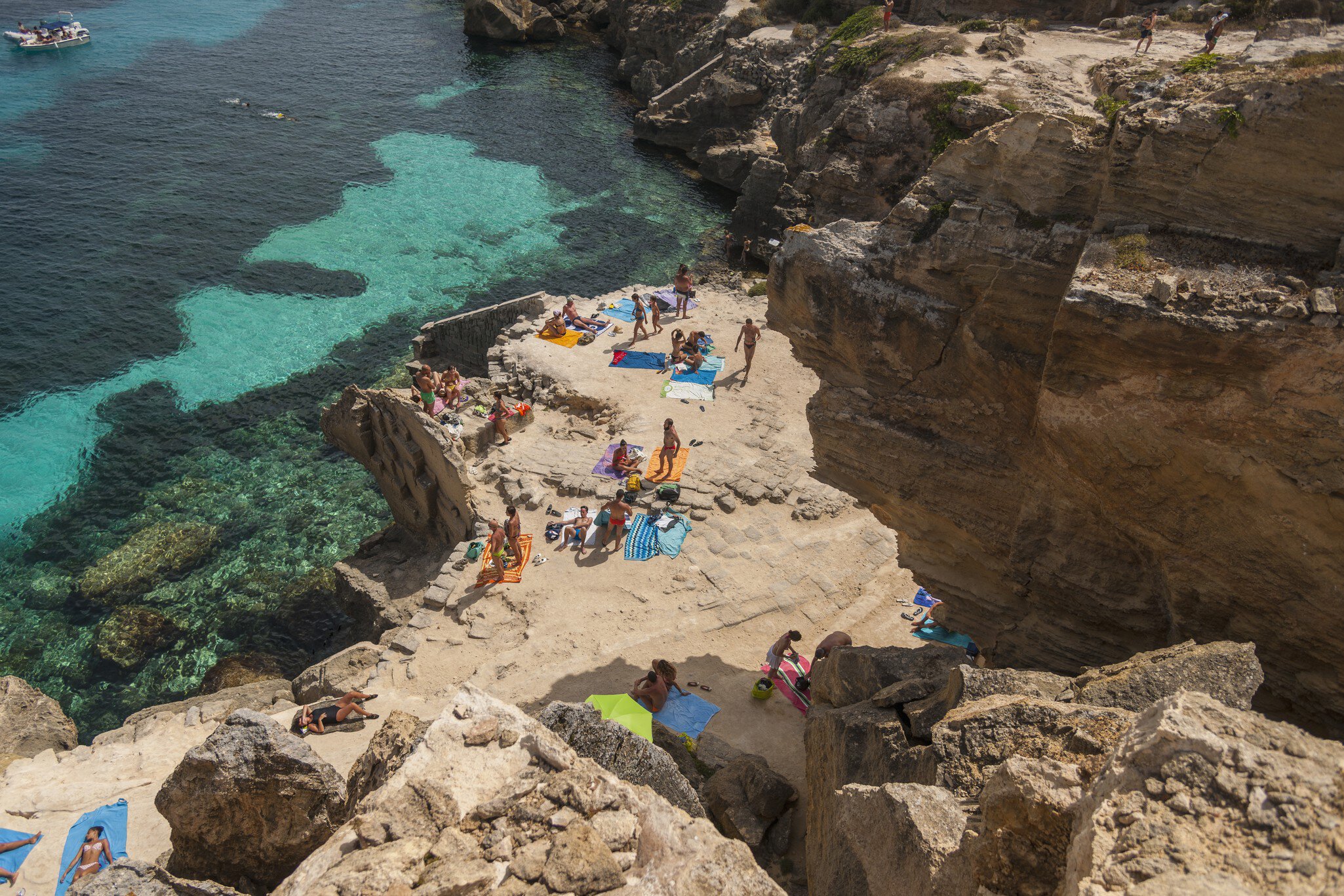 Aerial view of people sunbathing among rocks in a cove beach with clear ocean water beyond on a sunny day.