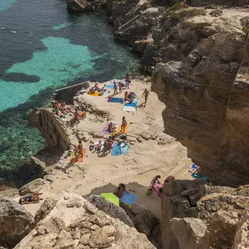 Aerial view of people sunbathing among rocks in a cove beach with clear ocean water beyond on a sunny day.