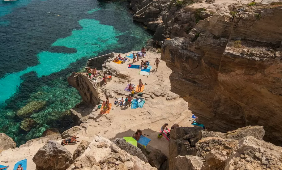 Aerial view of people sunbathing among rocks in a cove beach with clear ocean water beyond on a sunny day in Favignana